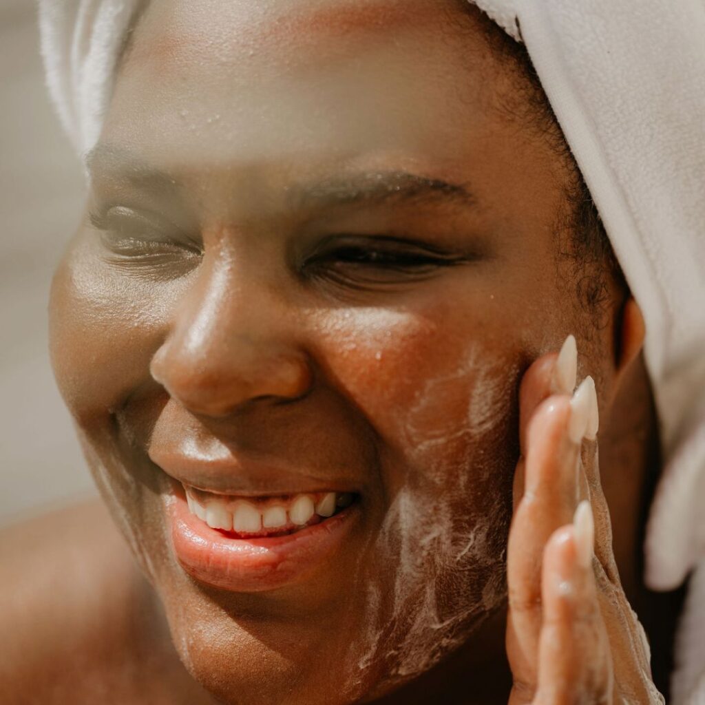A smiling woman fresh out of the shower washes her face with soap, presumably made with essential oils. 