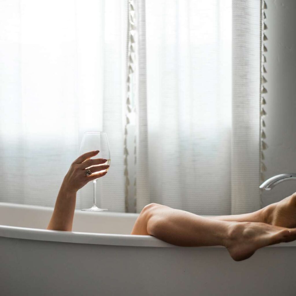 a woman is relaxing in the bathtub with a glass of wine, her legs dangling. 