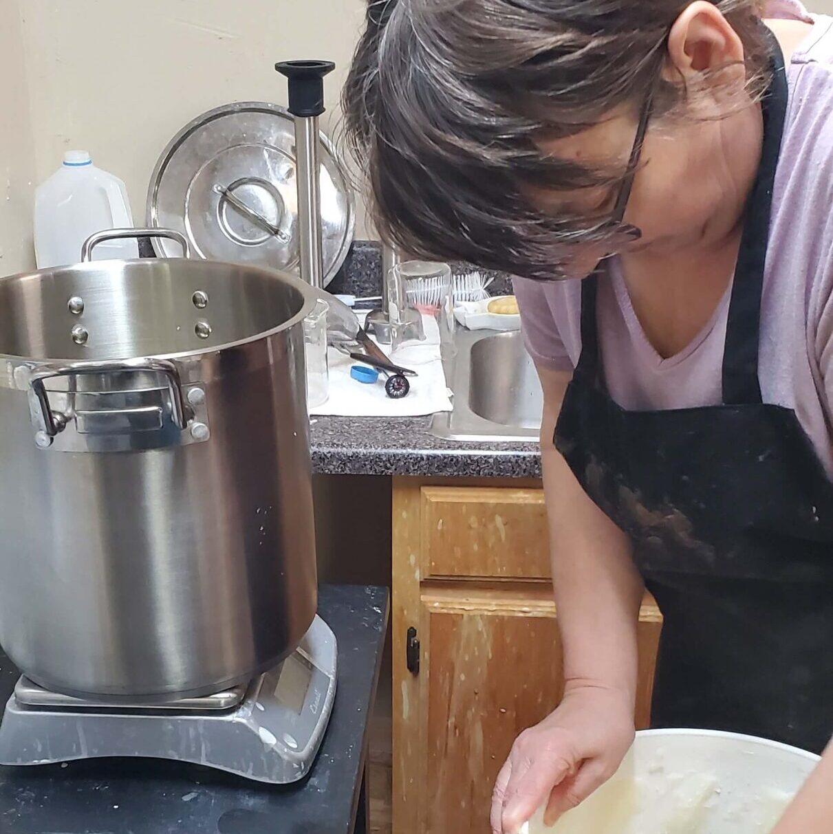 Susan making natural soap. A large cooking pot rests on a scale. Susan is working soap in a bucket. 
